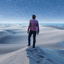 A man standing with his back to the camera, on a sand dune in the dessert, looking off in to the distance at a star filled sky